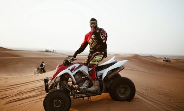 A male rider on an ATV navigating the sand dunes during the day, wearing protective gear and a helmet.